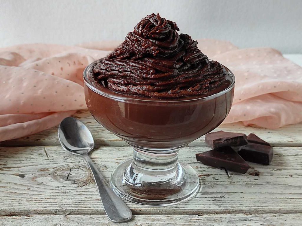 Crema di burro al cioccolato montata e vellutata, servita in coppa di vetro con cioccolato fondente a pezzi.