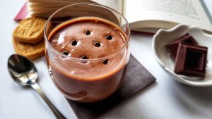 Un bicchiere di vetro con crema al cioccolato, su un tavolo accanto a dei biscotti e dei pezzi di cioccolato fondente