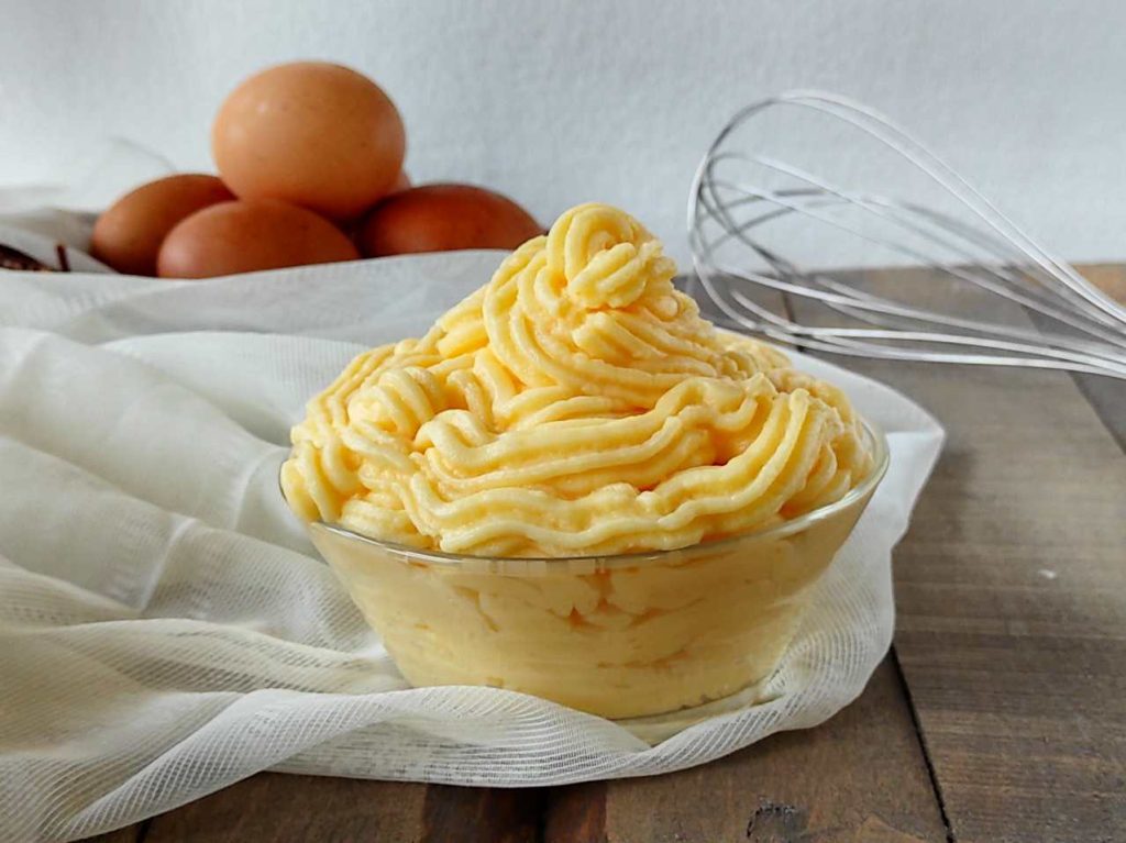 Crema pasticcera tradizionale decorata a ciuffi, servita in ciotola con uova e frusta sullo sfondo.